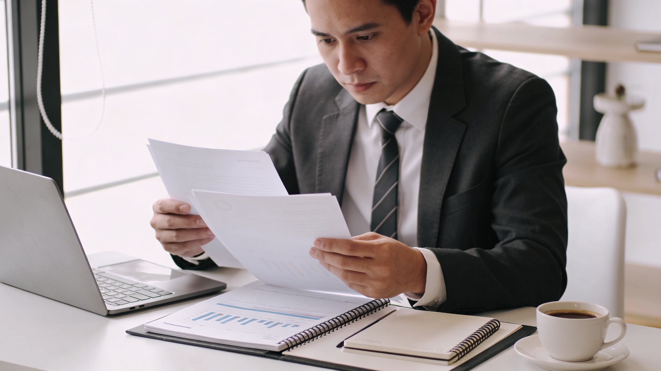 Un professionnel en costume examinant attentivement des documents financiers assis à son bureau dans un espace lumineux.
