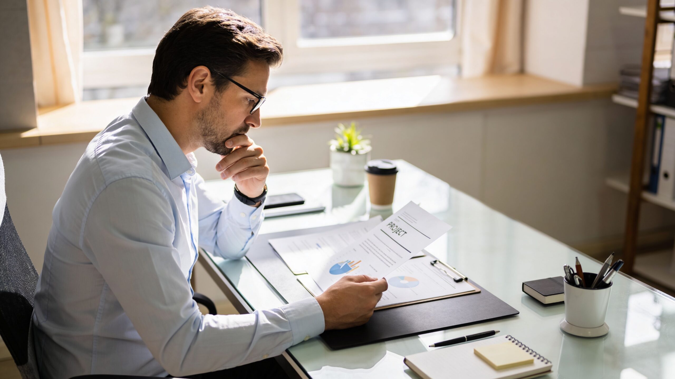 Un homme d'affaires concentré examinant des rapports de gestion de projet dans un bureau moderne et lumineux.